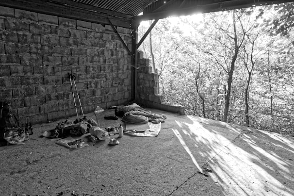 A black and white image depicting an area with improvised camping setup under a roofed structure, against a backdrop of partially visible trees. Camping gear such as a backpack, sleeping bag, trekking poles, and various items are spread on a flat concrete surface. The sunlight filters through the trees, casting shadows on the ground.