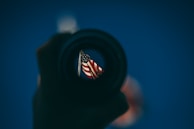 An American flag is viewed through the lens of a camera or telescope, with a background of a clear blue sky. The image is framed by the dark, round outline of the lens, creating a focused view of the flag.