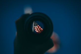 An American flag is viewed through the lens of a camera or telescope, with a background of a clear blue sky. The image is framed by the dark, round outline of the lens, creating a focused view of the flag.
