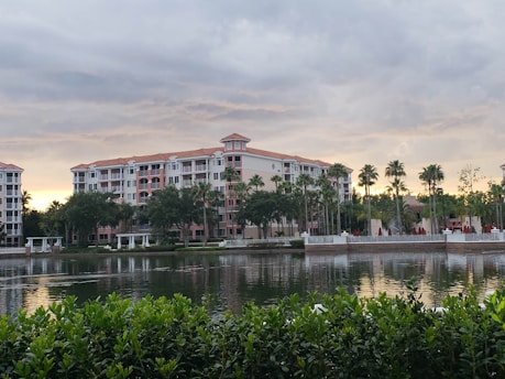 A large multi-story residential building with terracotta roof tiles is surrounded by lush greenery and palm trees. In the foreground, a calm body of water reflects the building's image, and shrubbery lines the bottom edge of the picture. The sky is overcast with a hint of sunlight breaking through the clouds.