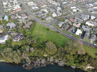 An aerial view of a suburban area featuring a network of roads, residential buildings, and a green park with trees. The landscape includes a river or lake at the bottom edge, bordered by dense foliage.