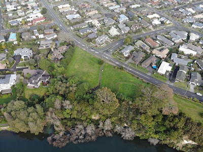 An aerial view of a suburban area featuring a network of roads, residential buildings, and a green park with trees. The landscape includes a river or lake at the bottom edge, bordered by dense foliage.