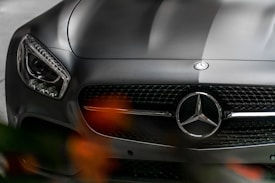 Close-up of a luxury car's front, showcasing the emblem prominently centered above a sleek, glossy grille with detailed design. The left headlight is visible, offering a modern and polished appearance, while the lighting and shadows highlight the car's streamlined, elegant curves. Blurred orange-red lights appear at the bottom, adding depth and contrast to the scene.