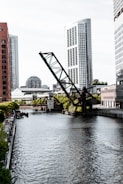 A river runs through an urban landscape bordered by modern skyscrapers. A large, angled drawbridge dominates the scene, partially raised above the water. The buildings, reflecting the light, exhibit sleek, contemporary architecture. Trees and smaller buildings line the banks, adding a touch of greenery to the city environment.