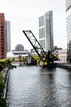 A river runs through an urban landscape bordered by modern skyscrapers. A large, angled drawbridge dominates the scene, partially raised above the water. The buildings, reflecting the light, exhibit sleek, contemporary architecture. Trees and smaller buildings line the banks, adding a touch of greenery to the city environment.