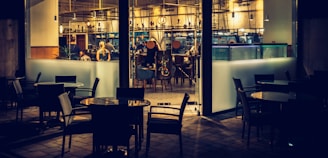 A dimly lit restaurant interior viewed from outside, with several empty tables and chairs in the foreground. Inside, a few people are seated at a counter and engaged in conversation. The decor features modern furnishings, warm lighting, and an array of hanging light fixtures.