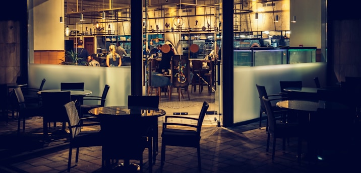 A dimly lit restaurant interior viewed from outside, with several empty tables and chairs in the foreground. Inside, a few people are seated at a counter and engaged in conversation. The decor features modern furnishings, warm lighting, and an array of hanging light fixtures.