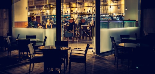 A dimly lit restaurant interior viewed from outside, with several empty tables and chairs in the foreground. Inside, a few people are seated at a counter and engaged in conversation. The decor features modern furnishings, warm lighting, and an array of hanging light fixtures.
