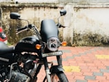 A black Royal Enfield motorcycle is parked beside a wall with some weathering and dirt marks. The vehicle has a prominent headlight, mirrors, and a visible license plate. The pavement is tiled in a pattern of red, yellow, and gray, and a portion of a car is seen to the left.
