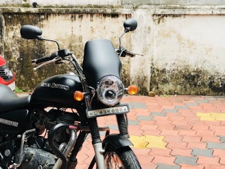 A black Royal Enfield motorcycle is parked beside a wall with some weathering and dirt marks. The vehicle has a prominent headlight, mirrors, and a visible license plate. The pavement is tiled in a pattern of red, yellow, and gray, and a portion of a car is seen to the left.