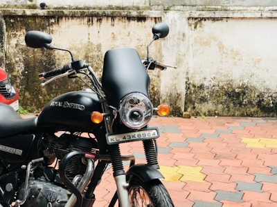 A black Royal Enfield motorcycle is parked beside a wall with some weathering and dirt marks. The vehicle has a prominent headlight, mirrors, and a visible license plate. The pavement is tiled in a pattern of red, yellow, and gray, and a portion of a car is seen to the left.