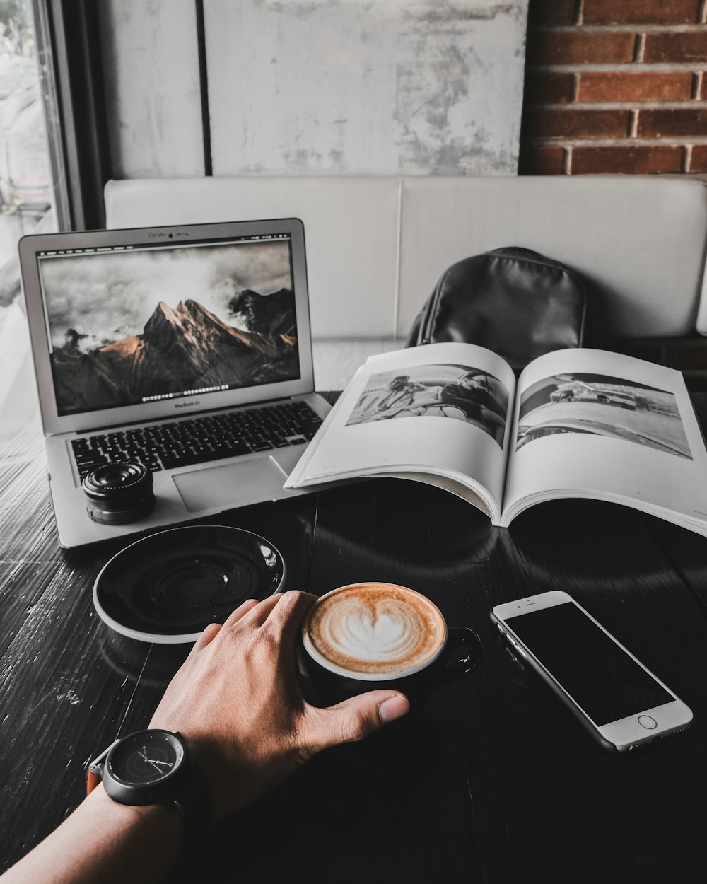 man holding cup of latte on table with MacBook Air and book