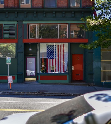 A storefront features a large American flag displayed in a window. The building facade includes dark green and brick red elements with a prominent red door on the right side. On the sidewalk, there are signs for parking and a small orange cone. Overhanging branches add a touch of nature to the otherwise urban scene.