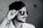 A black and white profile photograph of a person wearing dark sunglasses, tilting them slightly with their hand. The person has a short beard and mustache, with neatly styled hair. Their attire includes a light-colored, possibly patterned shirt, and there are several threads or bands wrapped around the wrist.
