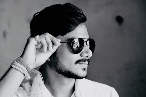 A black and white profile photograph of a person wearing dark sunglasses, tilting them slightly with their hand. The person has a short beard and mustache, with neatly styled hair. Their attire includes a light-colored, possibly patterned shirt, and there are several threads or bands wrapped around the wrist.