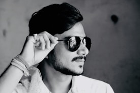 A black and white profile photograph of a person wearing dark sunglasses, tilting them slightly with their hand. The person has a short beard and mustache, with neatly styled hair. Their attire includes a light-colored, possibly patterned shirt, and there are several threads or bands wrapped around the wrist.
