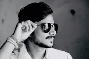 A black and white profile photograph of a person wearing dark sunglasses, tilting them slightly with their hand. The person has a short beard and mustache, with neatly styled hair. Their attire includes a light-colored, possibly patterned shirt, and there are several threads or bands wrapped around the wrist.