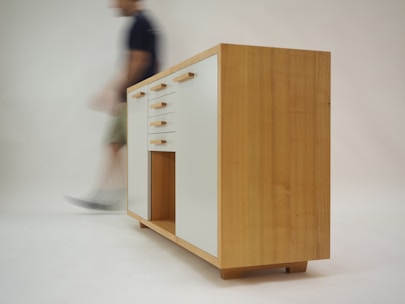 A wooden cabinet with modern design featuring white drawer fronts and wooden handles. The cabinet stands on short legs with a natural wood finish. A blurred figure of a person walks past in the background, creating a sense of motion.