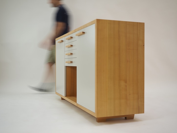 A wooden cabinet with modern design featuring white drawer fronts and wooden handles. The cabinet stands on short legs with a natural wood finish. A blurred figure of a person walks past in the background, creating a sense of motion.