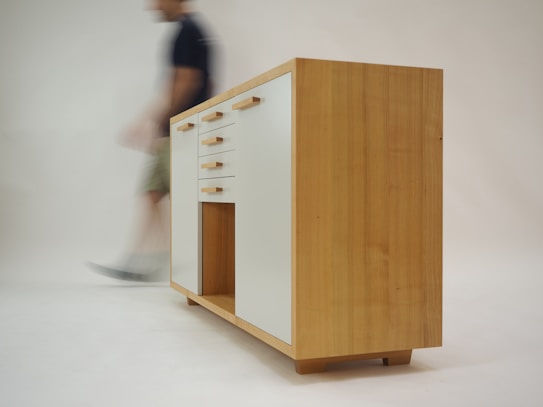 A wooden cabinet with modern design featuring white drawer fronts and wooden handles. The cabinet stands on short legs with a natural wood finish. A blurred figure of a person walks past in the background, creating a sense of motion.