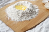 A wooden board holds a mound of flour with a well in the center containing a raw egg. Broken eggshells are visible to the side, and the setup suggests a preparation for making dough or pasta.