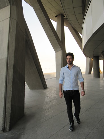 A man in a light blue shirt and dark pants walks confidently along a concrete pathway. The background features a modern architectural structure with large geometric pillars and an overhanging roof.
