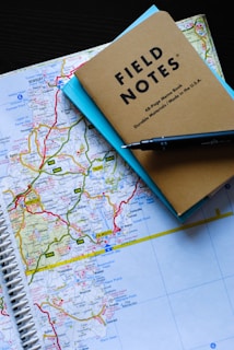 A spiral-bound map is laid out on a dark surface, featuring detailed geographical regions with roads highlighted in bright colors. Resting on top of the map are a brown notebook labeled 'Field Notes' and a light blue paper underneath. A black pen is diagonally placed across the top right corner of the notebook.