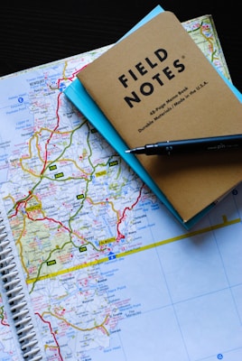 A spiral-bound map is laid out on a dark surface, featuring detailed geographical regions with roads highlighted in bright colors. Resting on top of the map are a brown notebook labeled 'Field Notes' and a light blue paper underneath. A black pen is diagonally placed across the top right corner of the notebook.