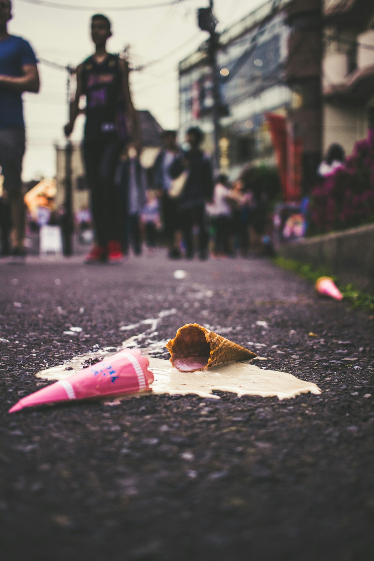 a melted ice cream cone on the asphalt.  It has a pink wrapper, skinny side pointing left and the cone doing the same pointing right. There is a crowd of blurred people milling around the in the blurred city. 