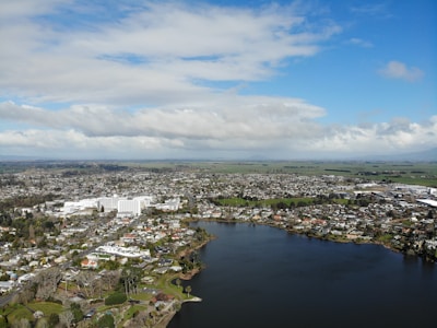 An aerial view of a suburban area featuring a large body of water in the foreground. Numerous houses and buildings are spread out across the landscape with a mix of green spaces and trees. The sky is partly cloudy with patches of blue, and the horizon shows distant fields and more scattered development.