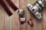 A vintage silver camera with a lens is placed on a wooden surface, surrounded by various types of film rolls. Two strips of developed negatives are positioned to the left, while two film canisters and a black and white film box sit to the right.