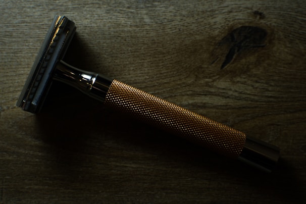 A metal safety razor is placed on a wooden surface. The razor has a shiny, metallic finish with a textured grip handle.