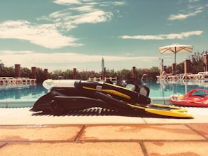 Snorkeling gear is placed on the edge of a swimming pool, including fins, a mask, and a snorkel. Sun loungers are visible in the background, alongside people relaxing under large umbrellas. The sky is bright with scattered clouds, and the scene has a tranquil vacation vibe.