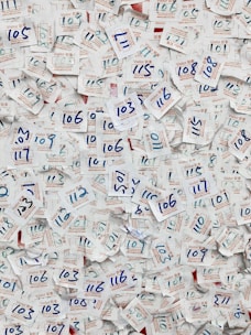 A large collection of visitor badges with handwritten numbers scattered chaotically. The numbers are mainly in blue ink, and the badges are made of paper with both English and Chinese text printed in red and blue. The background is mostly paper, displaying a dense and overlapping pattern due to the number of badges.