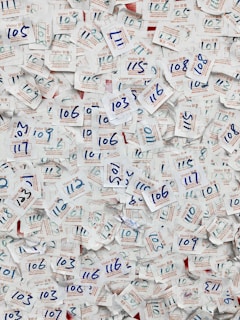 A large collection of visitor badges with handwritten numbers scattered chaotically. The numbers are mainly in blue ink, and the badges are made of paper with both English and Chinese text printed in red and blue. The background is mostly paper, displaying a dense and overlapping pattern due to the number of badges.