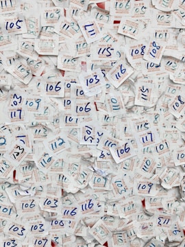 A large collection of visitor badges with handwritten numbers scattered chaotically. The numbers are mainly in blue ink, and the badges are made of paper with both English and Chinese text printed in red and blue. The background is mostly paper, displaying a dense and overlapping pattern due to the number of badges.