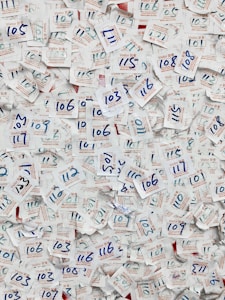A large collection of visitor badges with handwritten numbers scattered chaotically. The numbers are mainly in blue ink, and the badges are made of paper with both English and Chinese text printed in red and blue. The background is mostly paper, displaying a dense and overlapping pattern due to the number of badges.