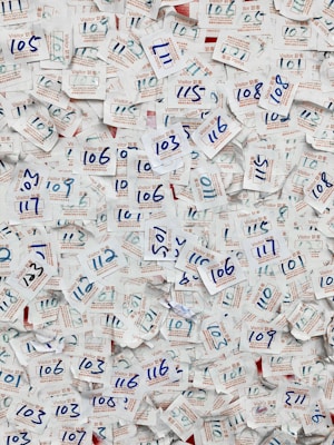 A large collection of visitor badges with handwritten numbers scattered chaotically. The numbers are mainly in blue ink, and the badges are made of paper with both English and Chinese text printed in red and blue. The background is mostly paper, displaying a dense and overlapping pattern due to the number of badges.