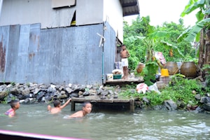 A group of children are swimming in a narrow waterway next to a rustic shack made of metal sheets. The area is surrounded by lush green banana plants and other vegetation. There are various containers and items on the ground near the shack, including a yellow bucket and large jars. A man in white shorts stands on a platform by the water.