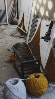 A room contains large rug canvases leaning against a wall, partially completed with geometric patterns in black and brown. Spools of yarn in various colors, including gray and mustard yellow, are placed on the floor next to the weaving equipment. The space has a somewhat unfinished and creative atmosphere with scattered materials.