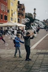 Children are playing with floating bubbles on a paved street surrounded by colorful buildings. Their excitement is evident as they reach out to touch the bubbles. The street is bustling with people, and there are streetlamps and cafe umbrellas in the background.