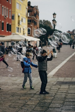 Children are playing with floating bubbles on a paved street surrounded by colorful buildings. Their excitement is evident as they reach out to touch the bubbles. The street is bustling with people, and there are streetlamps and cafe umbrellas in the background.
