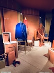 A fashion retail display features mannequins dressed in vibrant clothing. One mannequin wears a blue dress, while another is in an orange dress. Various accessories, such as handbags and shoes, are placed around the mannequins in a neatly arranged storefront. The backdrop includes paneling with artistic designs and muted lighting contributes to a chic ambiance.