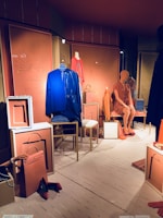 A fashion retail display features mannequins dressed in vibrant clothing. One mannequin wears a blue dress, while another is in an orange dress. Various accessories, such as handbags and shoes, are placed around the mannequins in a neatly arranged storefront. The backdrop includes paneling with artistic designs and muted lighting contributes to a chic ambiance.