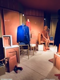 A fashion retail display features mannequins dressed in vibrant clothing. One mannequin wears a blue dress, while another is in an orange dress. Various accessories, such as handbags and shoes, are placed around the mannequins in a neatly arranged storefront. The backdrop includes paneling with artistic designs and muted lighting contributes to a chic ambiance.
