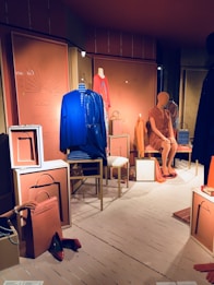 A fashion retail display features mannequins dressed in vibrant clothing. One mannequin wears a blue dress, while another is in an orange dress. Various accessories, such as handbags and shoes, are placed around the mannequins in a neatly arranged storefront. The backdrop includes paneling with artistic designs and muted lighting contributes to a chic ambiance.