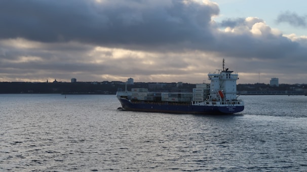A cargo ship sails across a calm body of water under a cloudy sky. The shoreline is visible in the background with some buildings and a tree line. The ship is carrying several containers stacked on its deck.
