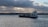 A cargo ship sails across a calm body of water under a cloudy sky. The shoreline is visible in the background with some buildings and a tree line. The ship is carrying several containers stacked on its deck.