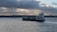 A cargo ship sails across a calm body of water under a cloudy sky. The shoreline is visible in the background with some buildings and a tree line. The ship is carrying several containers stacked on its deck.