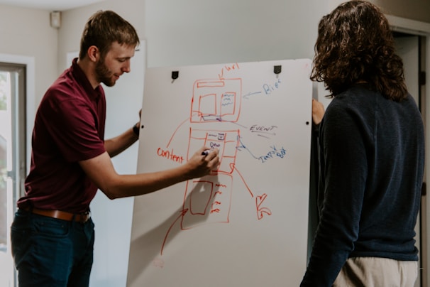 Two individuals are engaged in a discussion while standing near a whiteboard. One person is actively drawing with a marker, illustrating a diagram with boxes and arrows in red and blue ink. The other person observes attentively. Natural light filters through a nearby window, adding a soft glow to the environment.